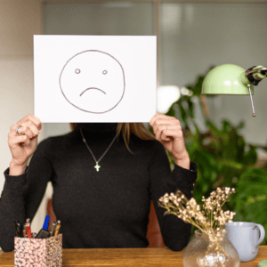 Une jeune femme tient une feuille devant son visage sur laquelle il y a un smiley triste.