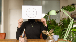 Une jeune femme tient une feuille devant son visage sur laquelle il y a un smiley triste.