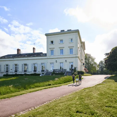 Manoir historique en pierre dans parc paysager avec pelouse, arbres et promeneurs.