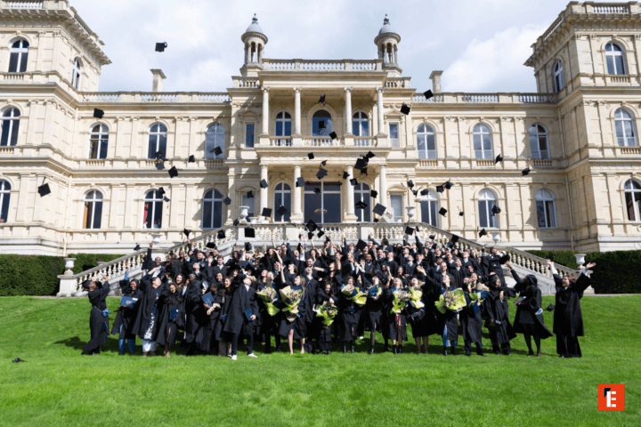 Cérémonie de remise de diplômes : diplômés en toques lançant leurs chapeaux devant bâtiment classique.