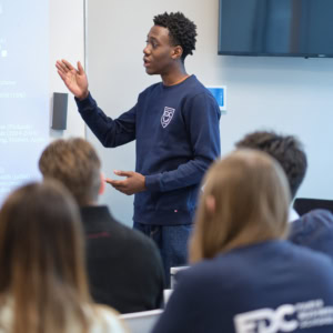 Formateur jeune présentant en classe moderne devant public attentif, écran et équipement audiovisuel.