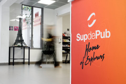 Réception SupdePub : couloir, panneau orange avec logo, miniature Tour Eiffel et personne floue.