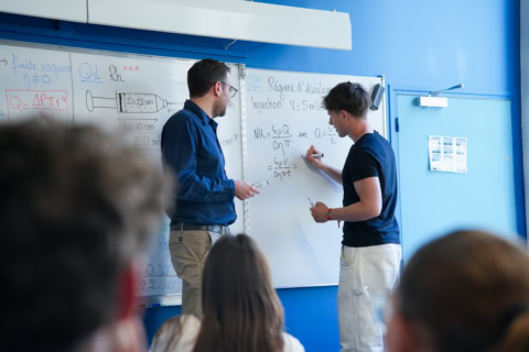 Cours collectif: professeur et étudiant écrivent au tableau blanc, élèves en arrière-plan.