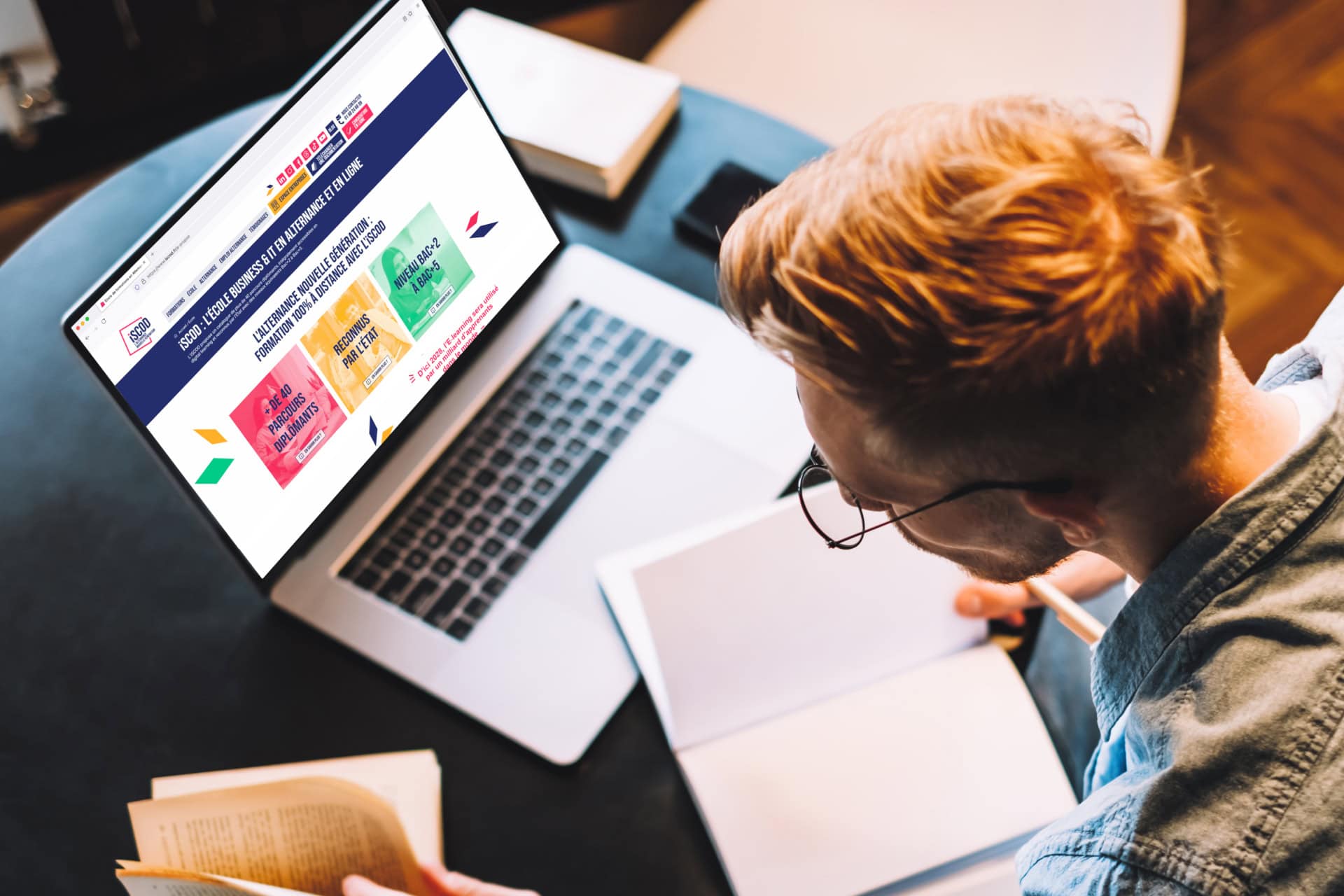 Étudiant prenant des notes avec ordinateur portable, carnet et livre sur bureau.