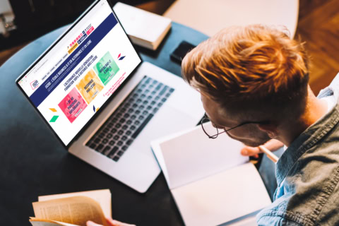 Étudiant prenant des notes avec ordinateur portable, carnet et livre sur bureau.