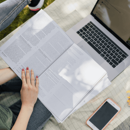 Lecture en plein air : personne en jean lisant un livre, ordinateur portable et smartphone.