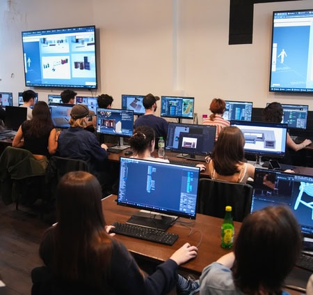 Salle informatique : étudiants concentrés sur ordinateurs et tablettes graphiques, atelier créatif nocturne.