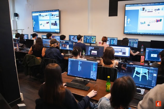 Salle informatique : étudiants concentrés sur ordinateurs et tablettes graphiques, atelier créatif nocturne.