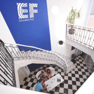 Atrium lumineux avec escalier courbe, logo Education First, carrelage damier et réunion.