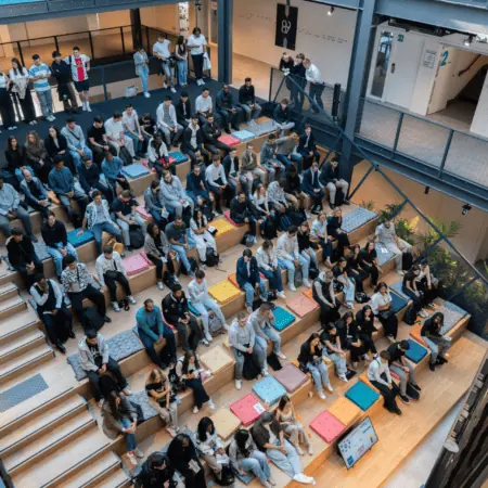 Atrium moderne avec gradins en bois, coussins colorés et public assis pour conférence.
