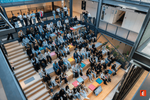 Atrium moderne avec gradins en bois, coussins colorés et public assis pour conférence.