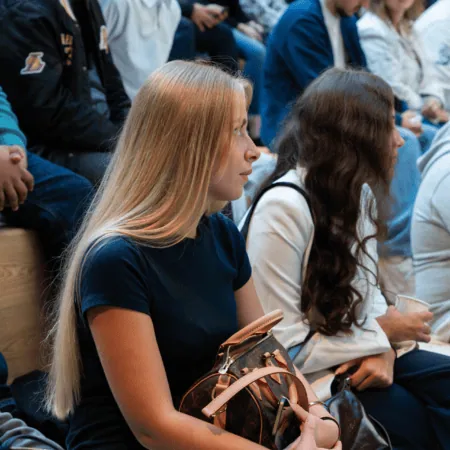 Femme blonde dans laudience assise sur des bancs, sac monogrammé.