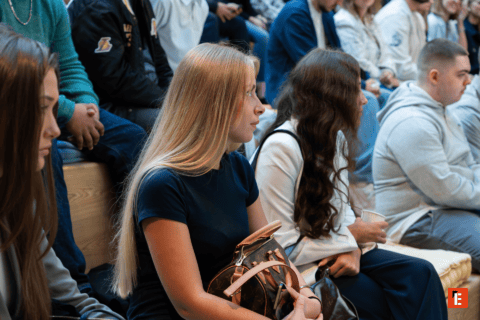 Femme blonde dans laudience assise sur des bancs, sac monogrammé.