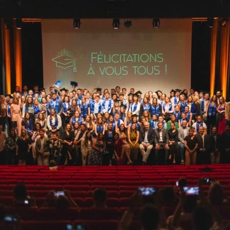 Cérémonie de remise de diplômes au théâtre, diplômés en écharpes bleues, public photographiant.