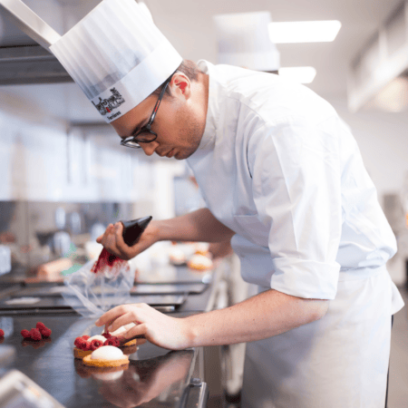 Chef pâtissier en toque dressant des tartelettes aux framboises en cuisine professionnelle.