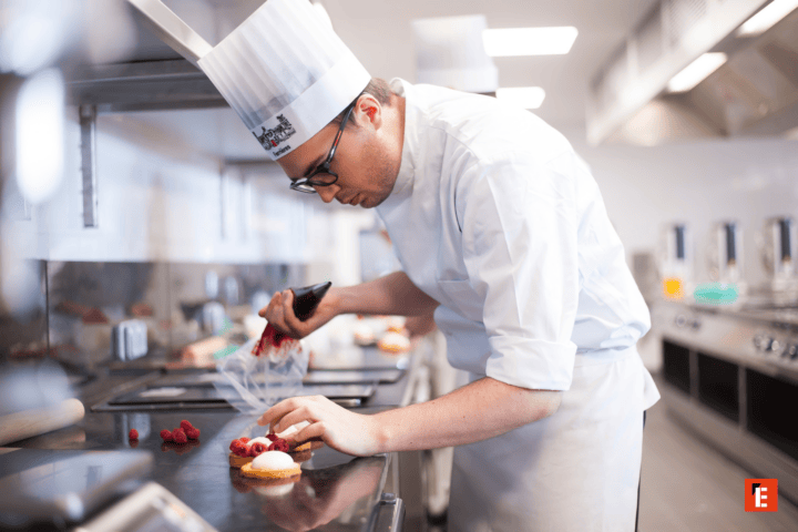 Chef pâtissier en toque dressant des tartelettes aux framboises en cuisine professionnelle.