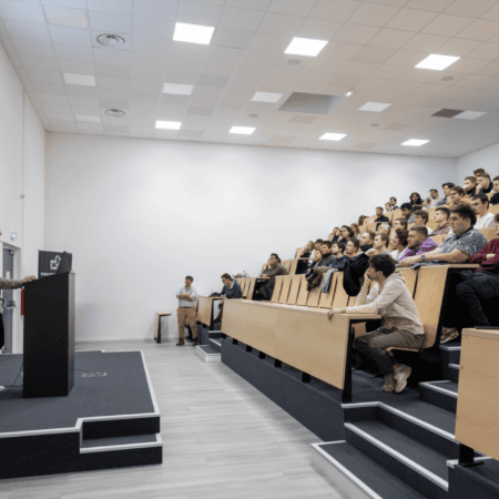 Conférencier devant amphithéâtre moderne rempli détudiants attentifs, projection de graphiques.