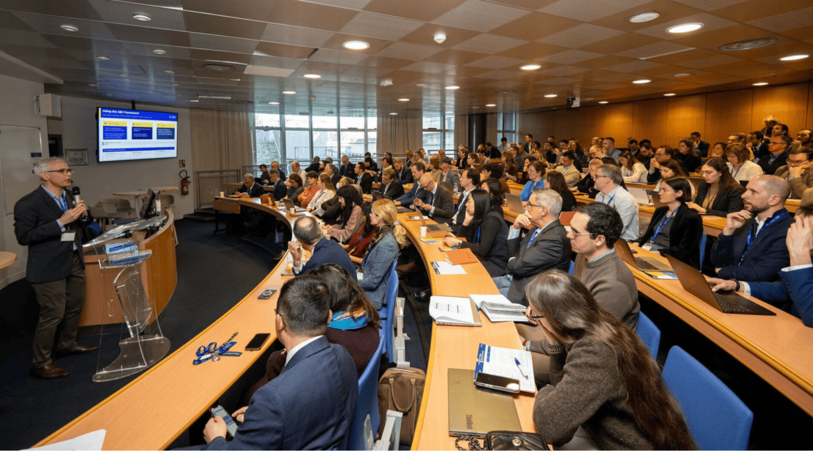 Conférence en amphithéâtre avec présentateur, diapositives projetées, public professionnel et ordinateurs.