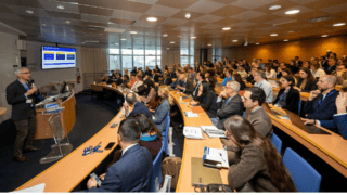 Conférence en amphithéâtre avec présentateur, diapositives projetées, public professionnel et ordinateurs.