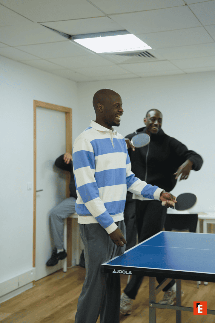 Deux hommes souriants jouent au ping-pong sur table JOOLA dans une salle commune détendue.