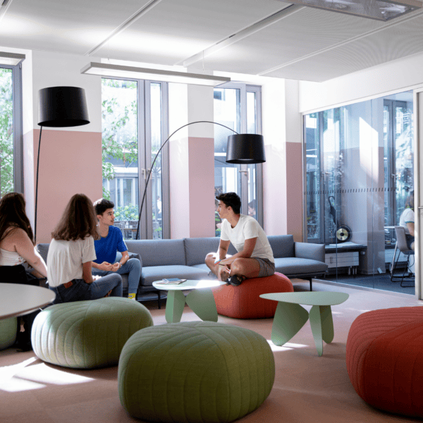 Espace détente scolaire lumineux avec baie vitrée, canapé, poufs et étudiants en discussion.