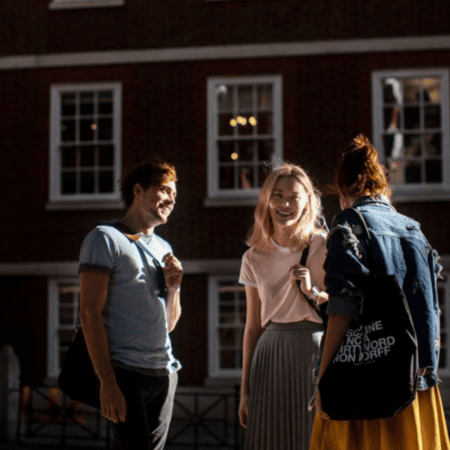 Étudiants discutant devant bâtiment en briques avec façade à fenêtres, scène urbaine ensoleillée.
