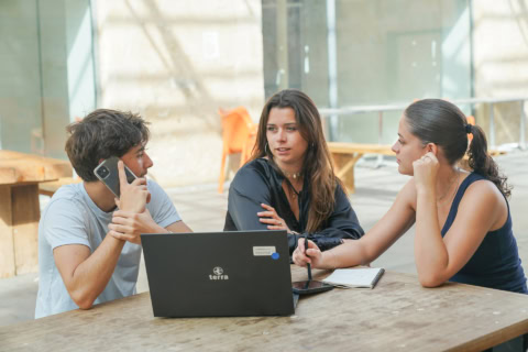 Trois étudiants en discussion autour dune table en bois, ordinateur portable et notes.