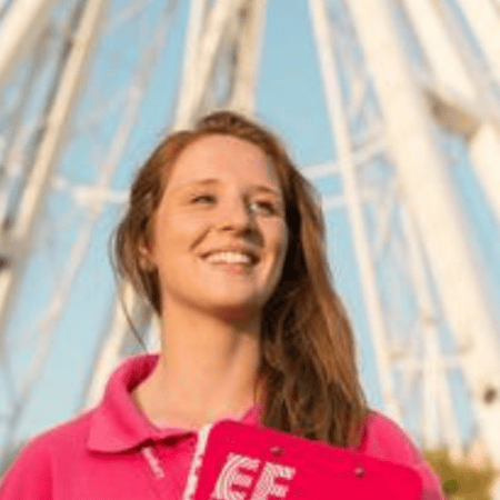 Femme daccueil en chemise rose, bloc-notes, souriante devant une grande roue ensoleillée.