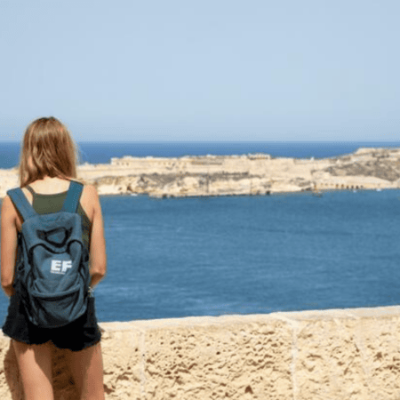 Femme de dos avec sac à dos bleu EF contemplant la mer et un bateau.