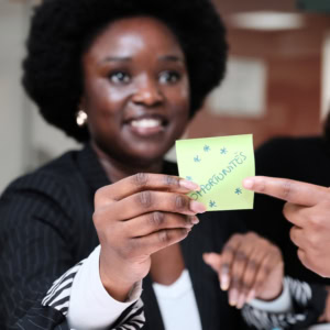 Femme afro souriante montrant un post-it vert «OPPORTUNITÉS» en brainstorming collaboratif au bureau.