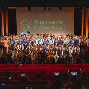 Remise de diplômes au théâtre : groupe détudiants en toge et stoles bleues, public applaudissant.