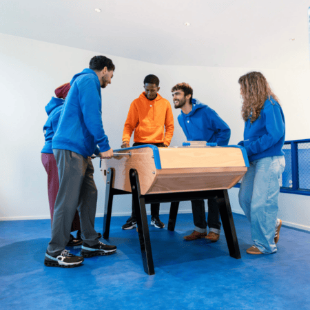 Groupe de joueurs autour dun baby-foot dans une salle de loisirs au sol bleu.