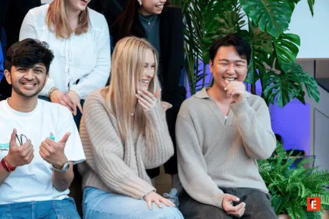 Groupe de jeunes adultes riant sur canapé, ambiance conviviale, plantes vertes en arrière-plan.