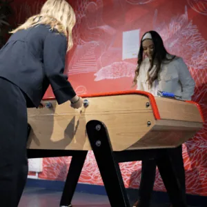 Joueurs de baby-foot sur table en bois, mural rouge, poignée bleue visible.