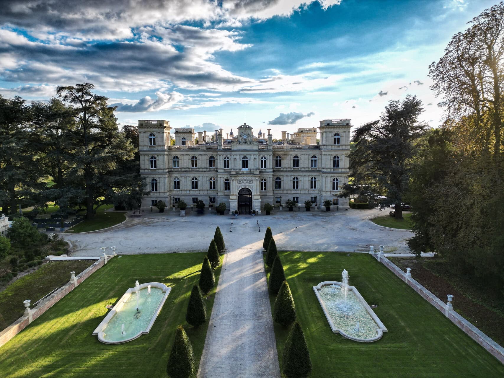 Manoir classique en pierre, allée centrée, bassins réfléchissants et fontaines dans jardins symétriques.