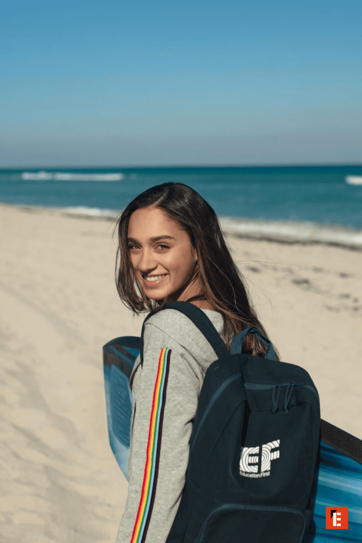 Jeune femme souriante à la plage avec sac à dos et planche de surf.