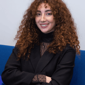 Portrait femme cheveux bouclés cuivrés souriante, blazer noir, dentelle, siège bleu.