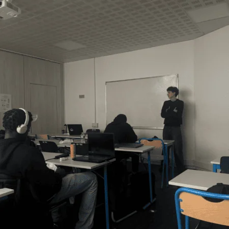 Salle de cours avec étudiants et ordinateurs portables, tableau blanc, casques et ambiance studieuse.