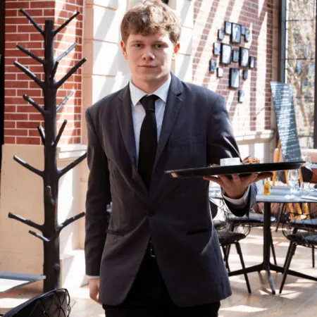 Serveur en costume portant un plateau avec café et croissant dans un café lumineux.