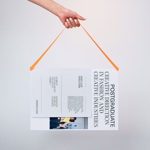 Affiche Mastère Direction Créative Mode ESMOD, brochure suspendue par main avec ruban orange.