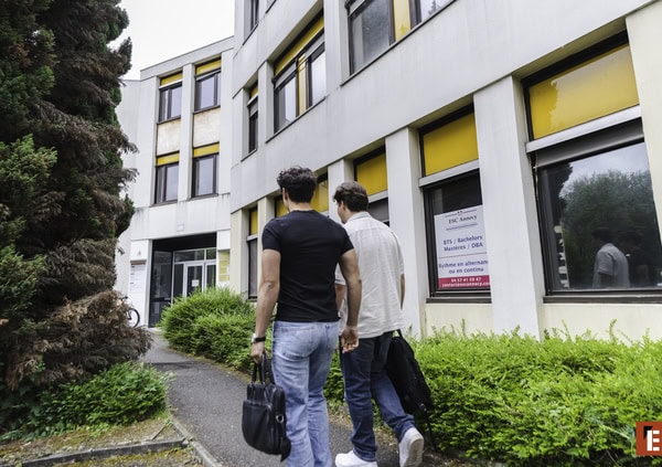 Bâtiment moderne à volets jaunes, façade avec fenêtres et deux hommes marchant vers lentrée.