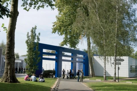Campus moderne, architecture bleue, arbres, pelouse et étudiants.