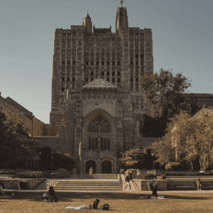 Entrée monumentale gothique du campus avec pelouse, escaliers et étudiants au soleil.