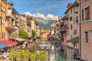Canal bordé de maisons pastel, pont en pierre, cafés en terrasse et montagnes en arrière-plan.