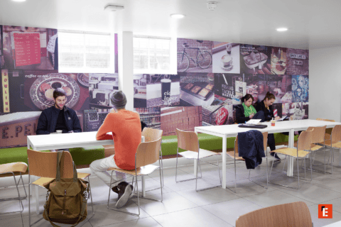 Espace commun moderne type café-étude avec grandes tables, chaises, étudiants et fresque murale.