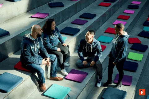 Étudiants HEC Paris en sweat à capuche discutant sur gradins damphithéâtre avec coussins colorés.