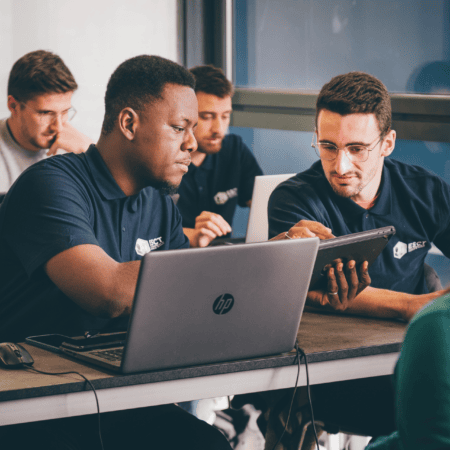 Hommes en formation professionnelle collaborant sur ordinateurs portables et tablettes en salle de classe.