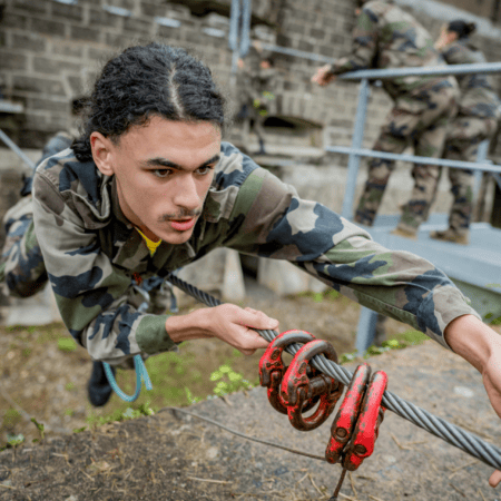 Jeune soldat en camouflage grimpe un câble dobstacle, harnais et poulies rouges visibles.