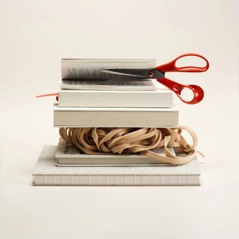 Nature morte minimaliste : ciseaux rouges sur pile de livres, ruban et carnets crème.