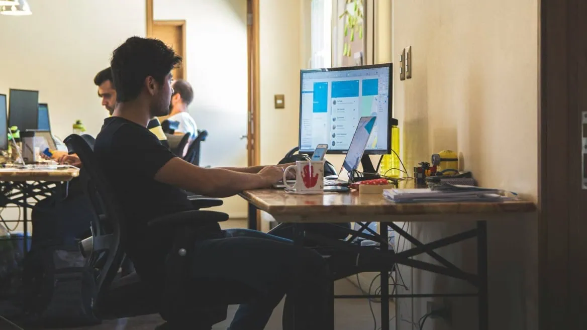 Open space bureau avec personne à lordinateur, écran, laptop, smartphone et accessoires de bureau.